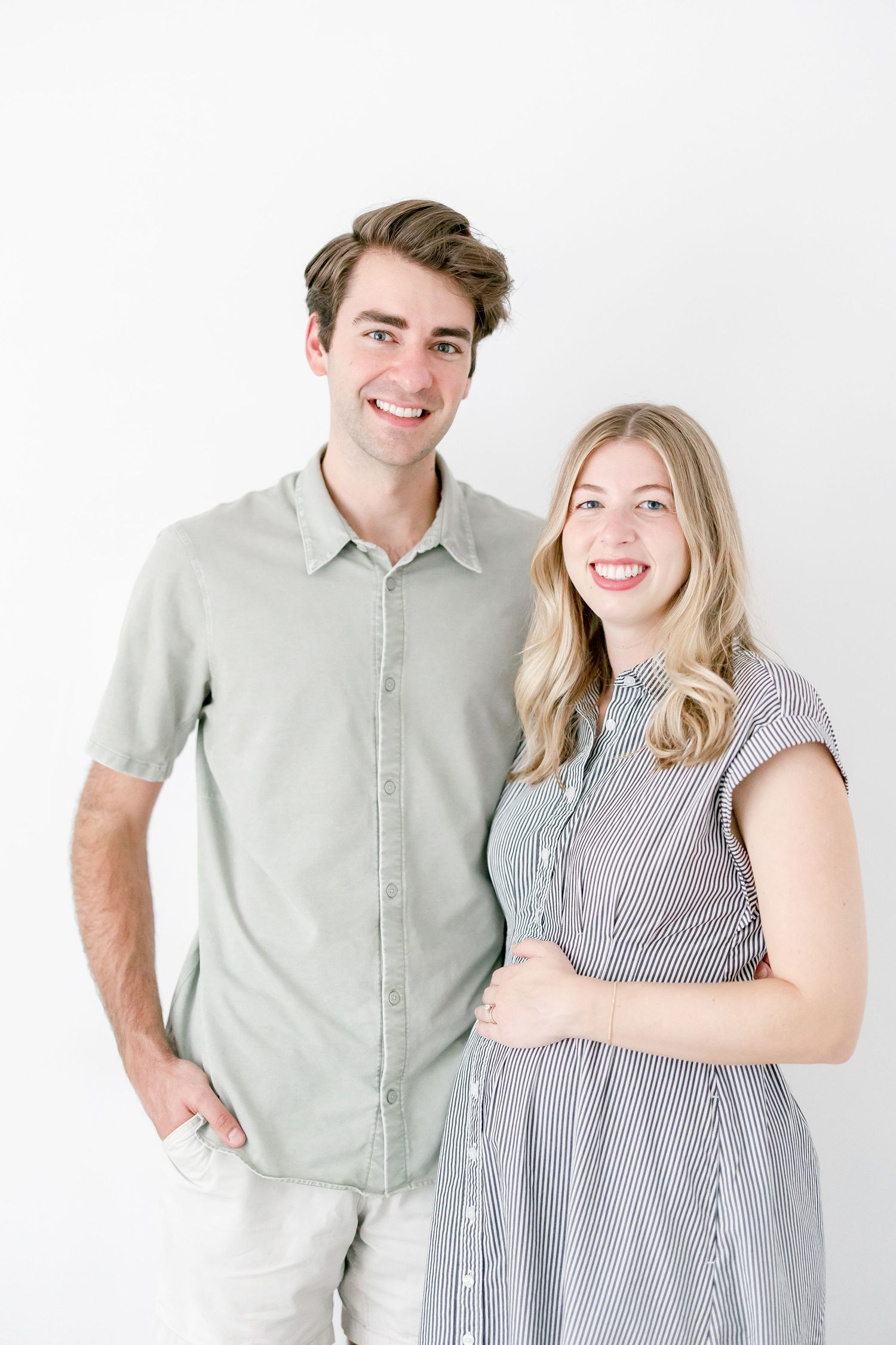 A couple in casual attire poses against a white background, wearing a sage green shirt and striped dress.