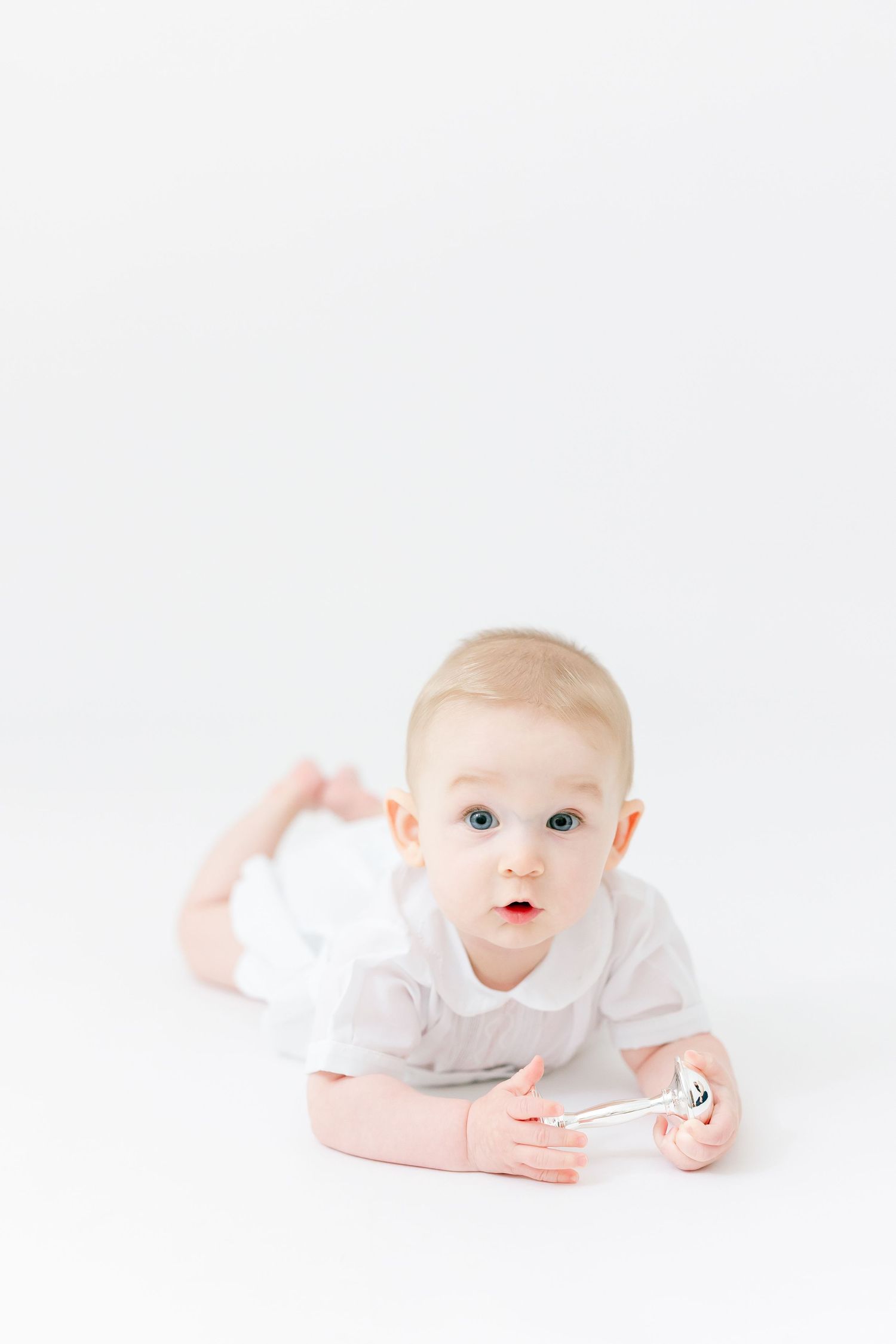 A cute baby in a white onesie lies on their tummy on a white background, holding a rattle and looking at the camera.
