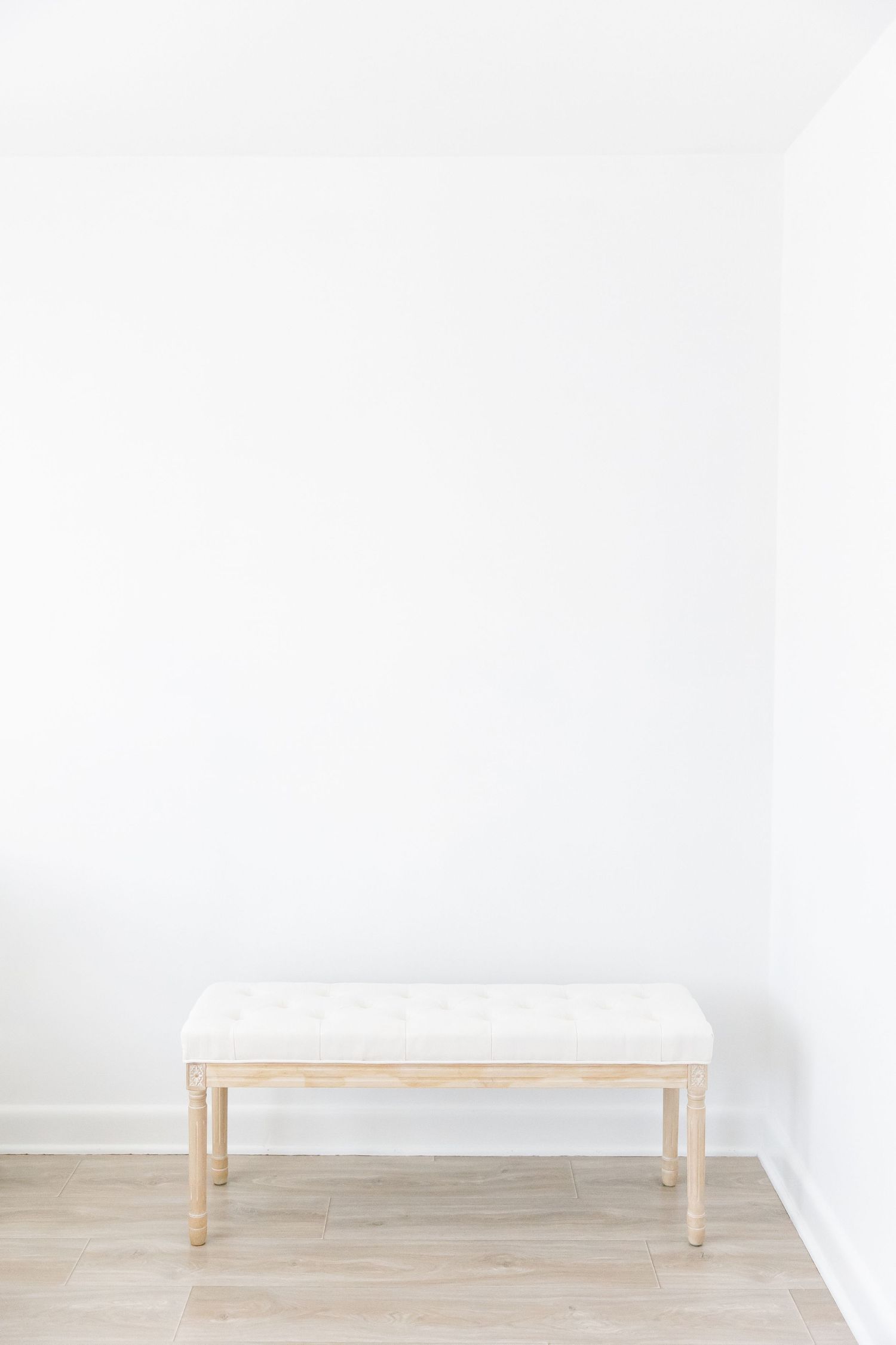 Minimalist white bench with wooden legs sits against blank white wall in bright empty room with light wood flooring.