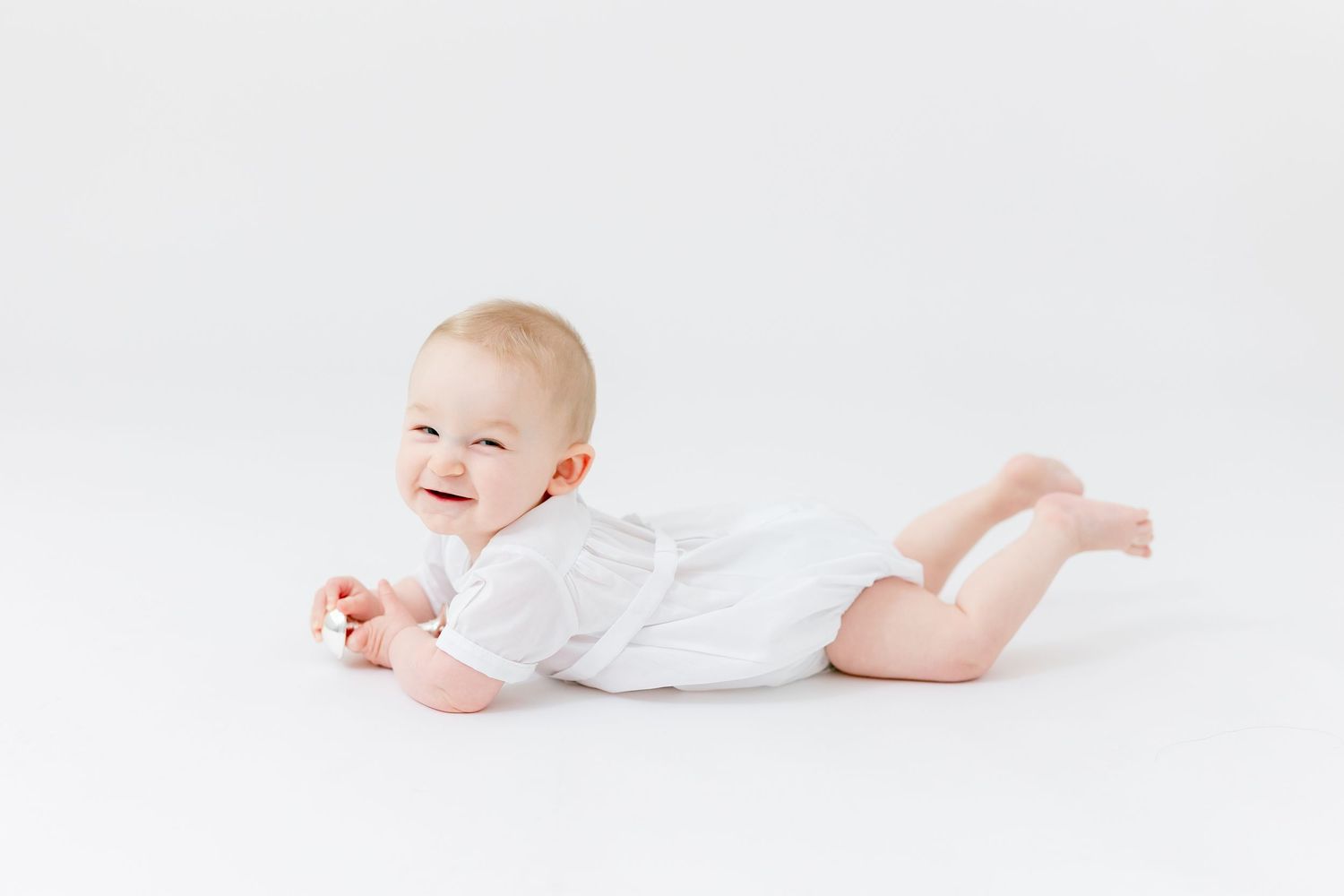 Smiling baby in a white outfit lying on their tummy and propping up on one arm against a clean, bright white studio background.