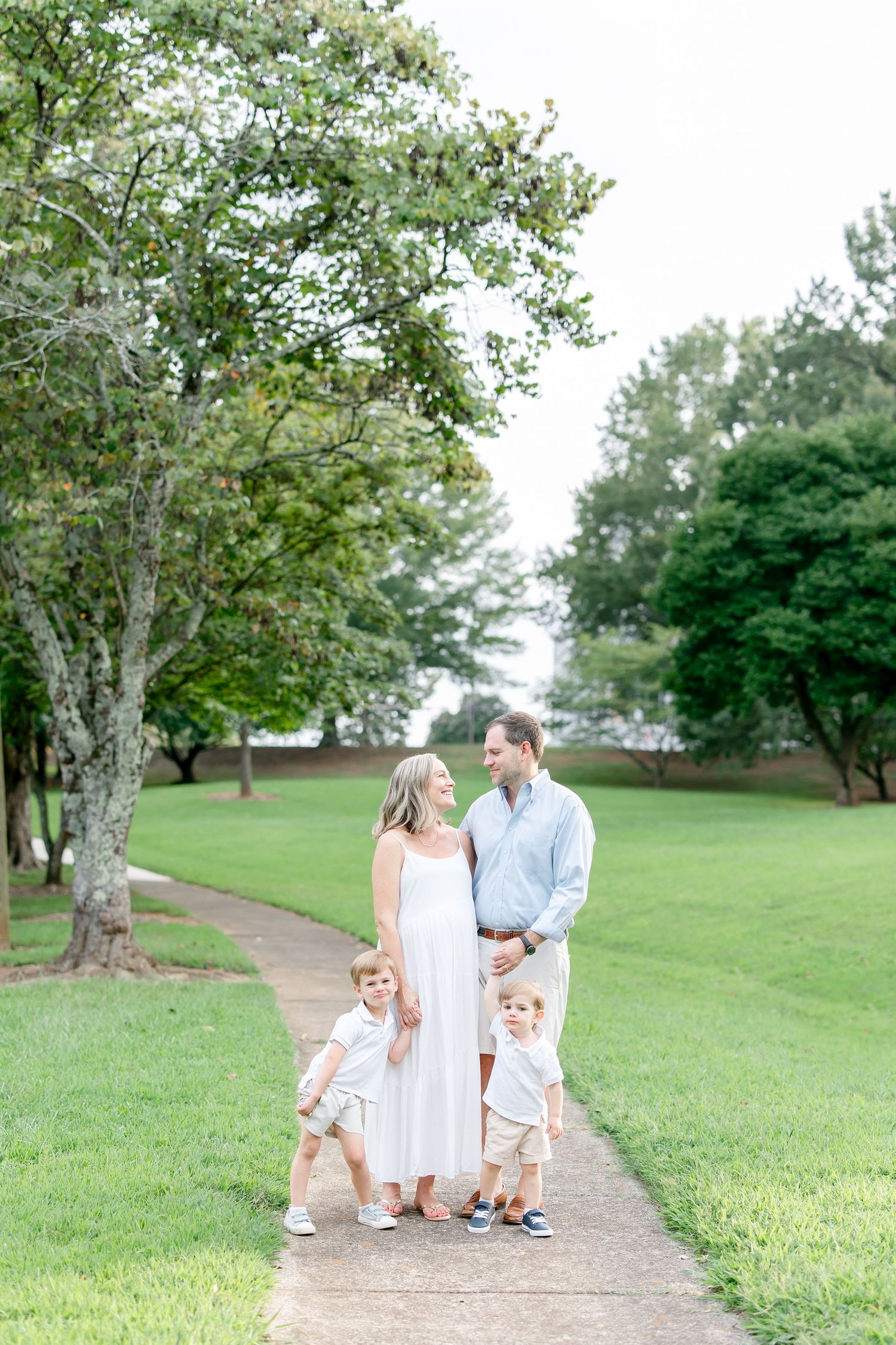 A family in white outfits walks together on a winding path through a green park with tall trees.