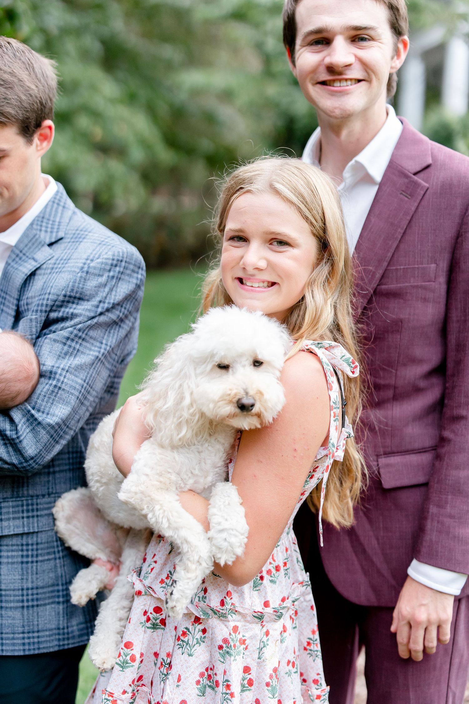 Small white fluffy dog being held during outdoor family gathering with people in dressy attire.