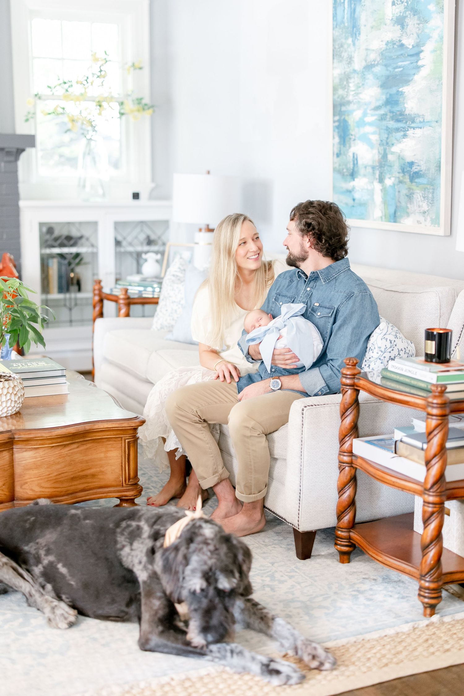Family relaxes on white armchair in bright living room with sleeping dog and blue artwork on wall.