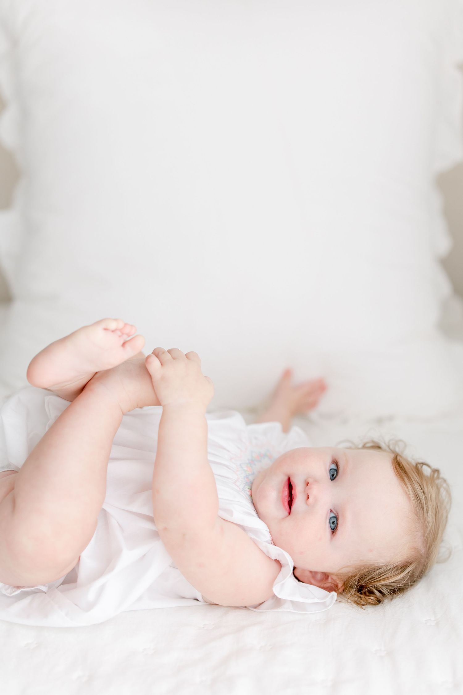 Baby in white dress lying on soft white bedding while playfully grabbing feet.
