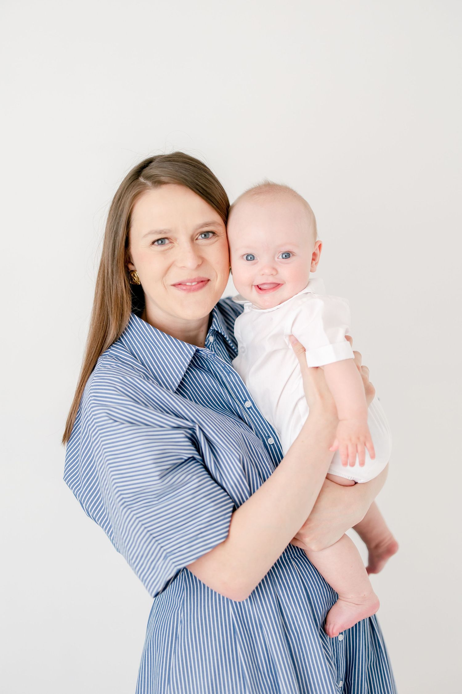 A parent in a striped blue dress holds a smiling baby wearing white against a neutral background.