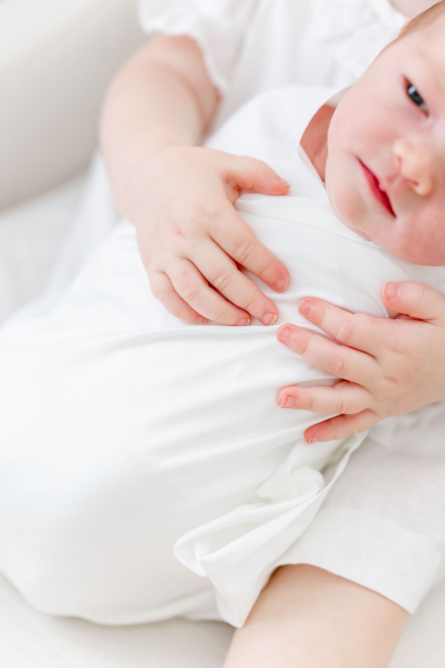 A baby wearing white clothing rests on white bedding while reaching out with small hands.