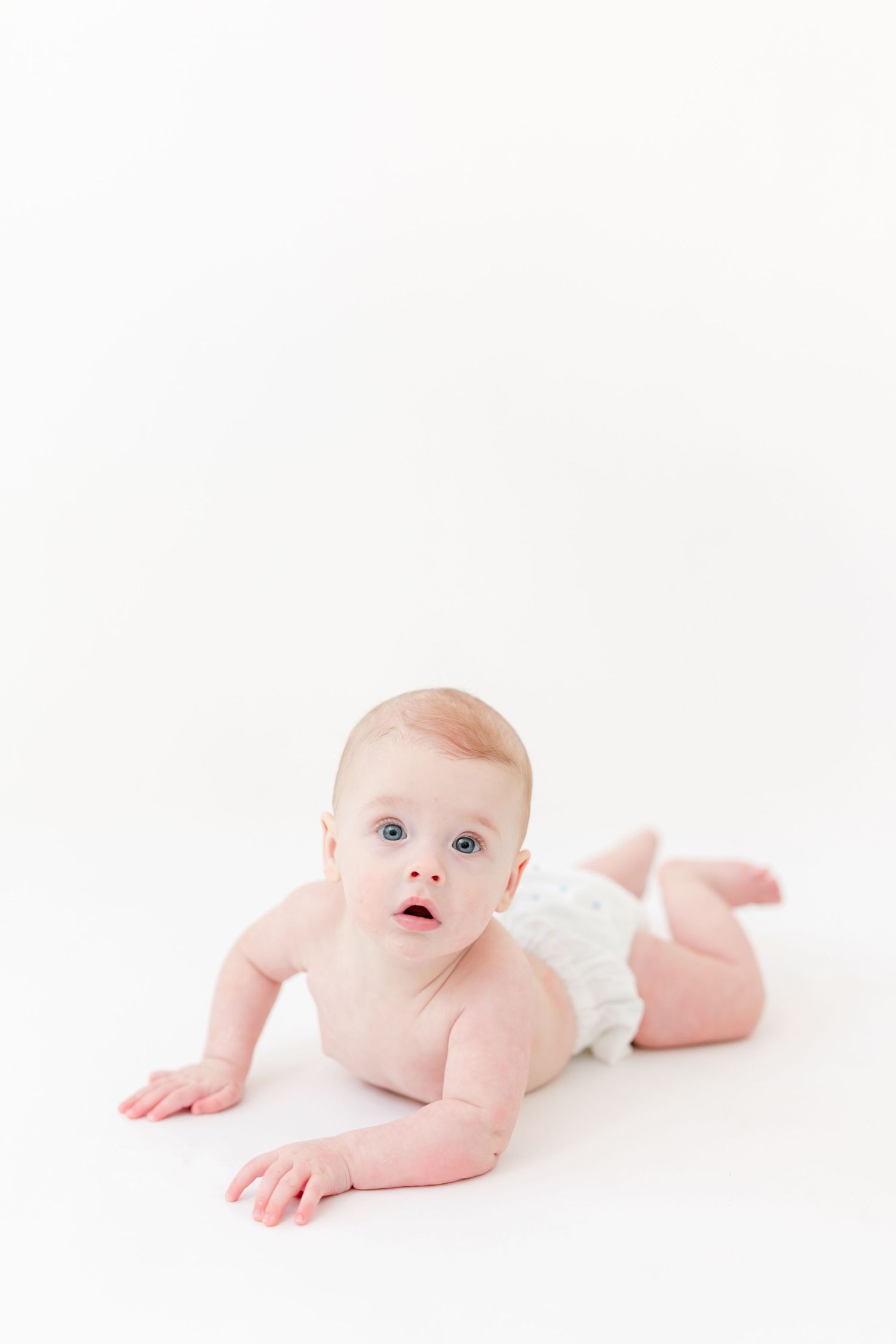 Baby with blue eyes lying on their tummy and lifting their head, wearing a white diaper against a bright white studio background.