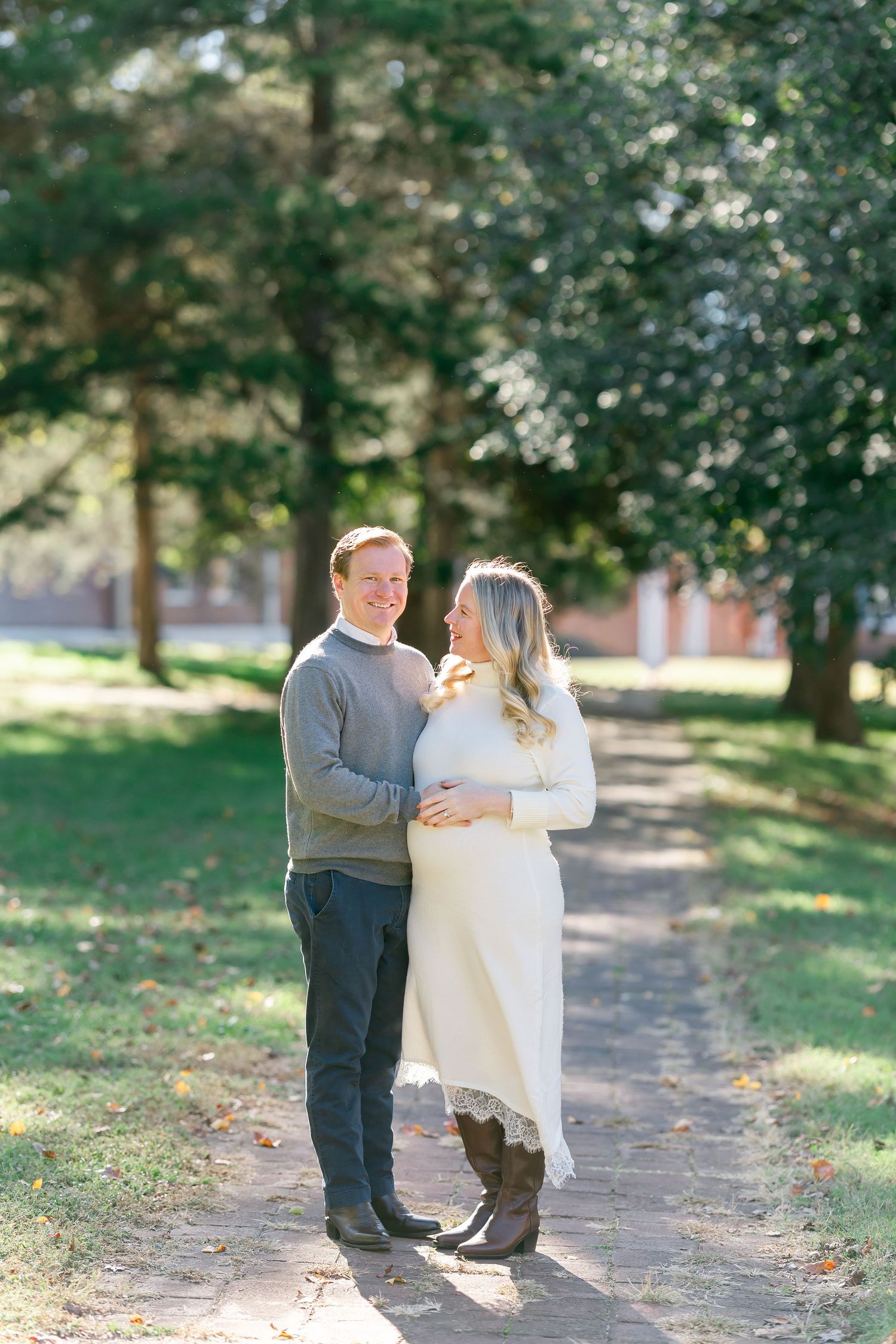 Smiling expectant couple embracing on a tree-lined park path during a sunny outdoor maternity photo session.