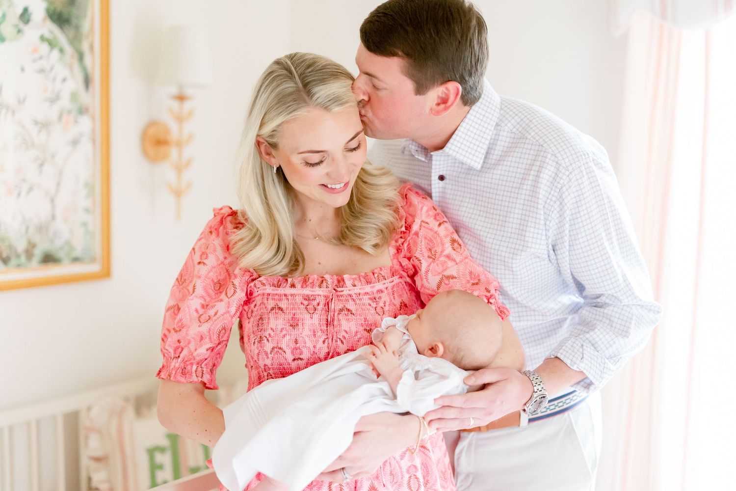 A couple smiles tenderly while holding their newborn baby, the mother wearing a pink floral dress in a softly lit nursery.