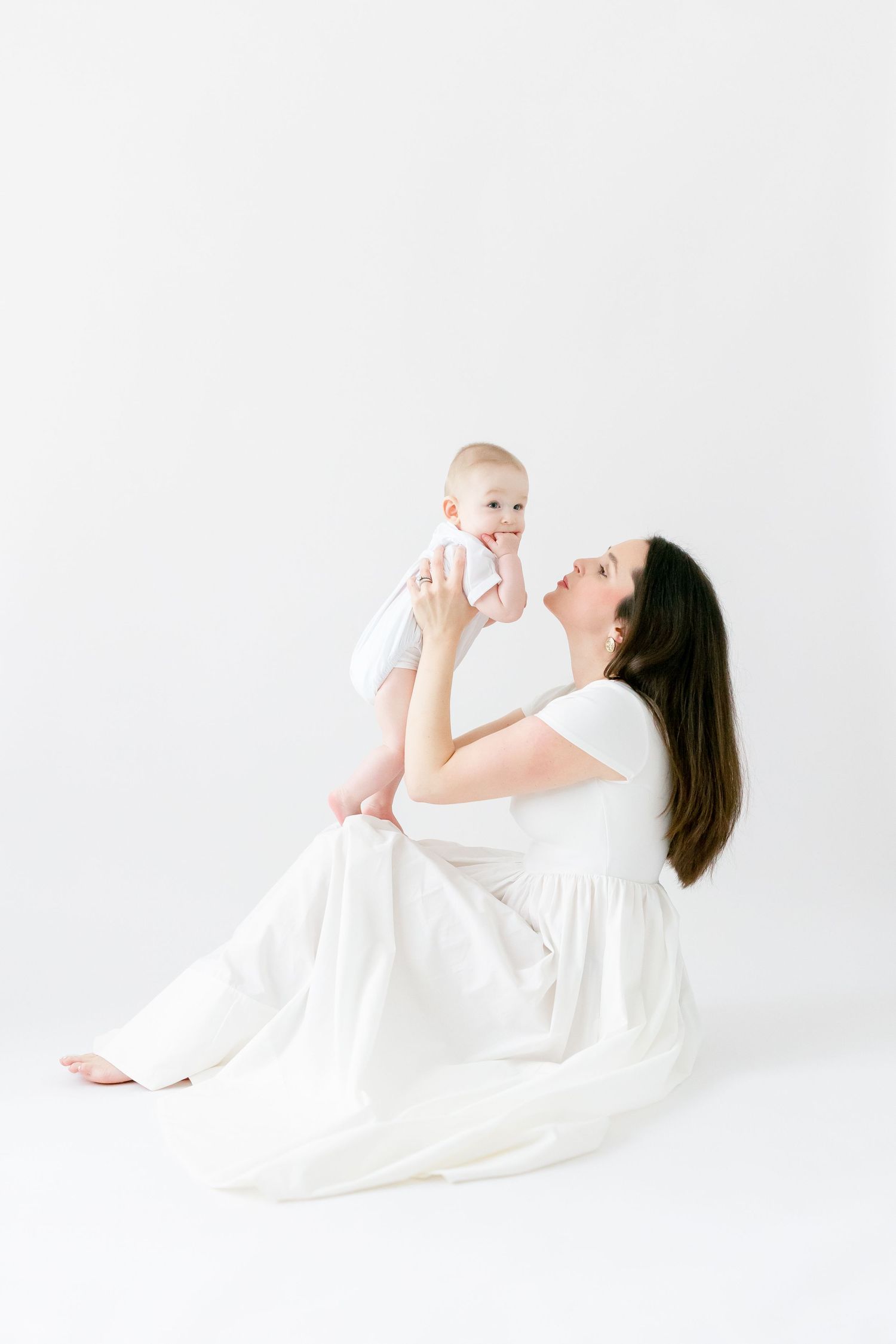 A mother in a white dress sits on the floor holding her smiling baby up in the air against a white background.