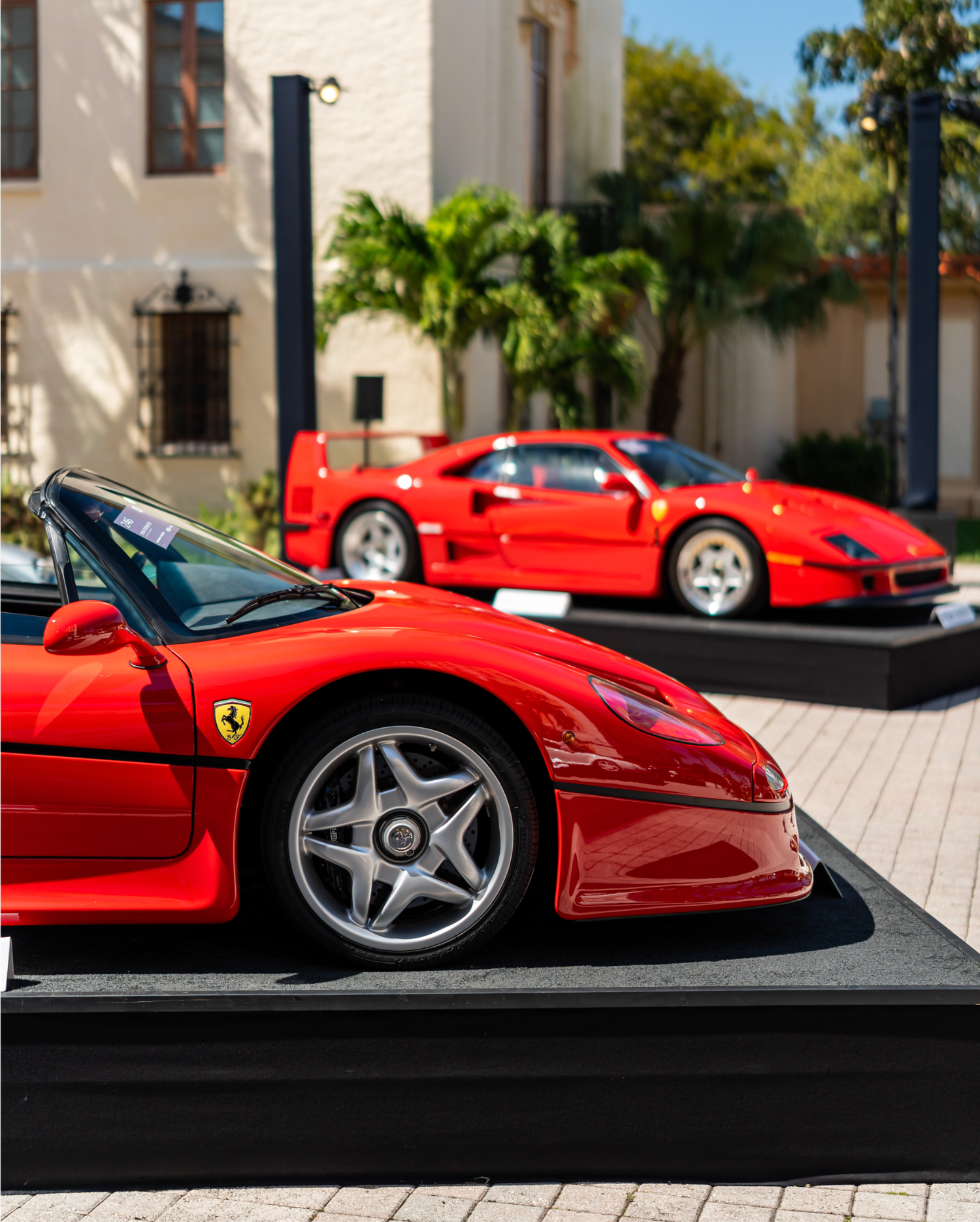 Red Ferrari F50 on display at a concours-style event, elevated on a platform with a red F40 visible in the background
