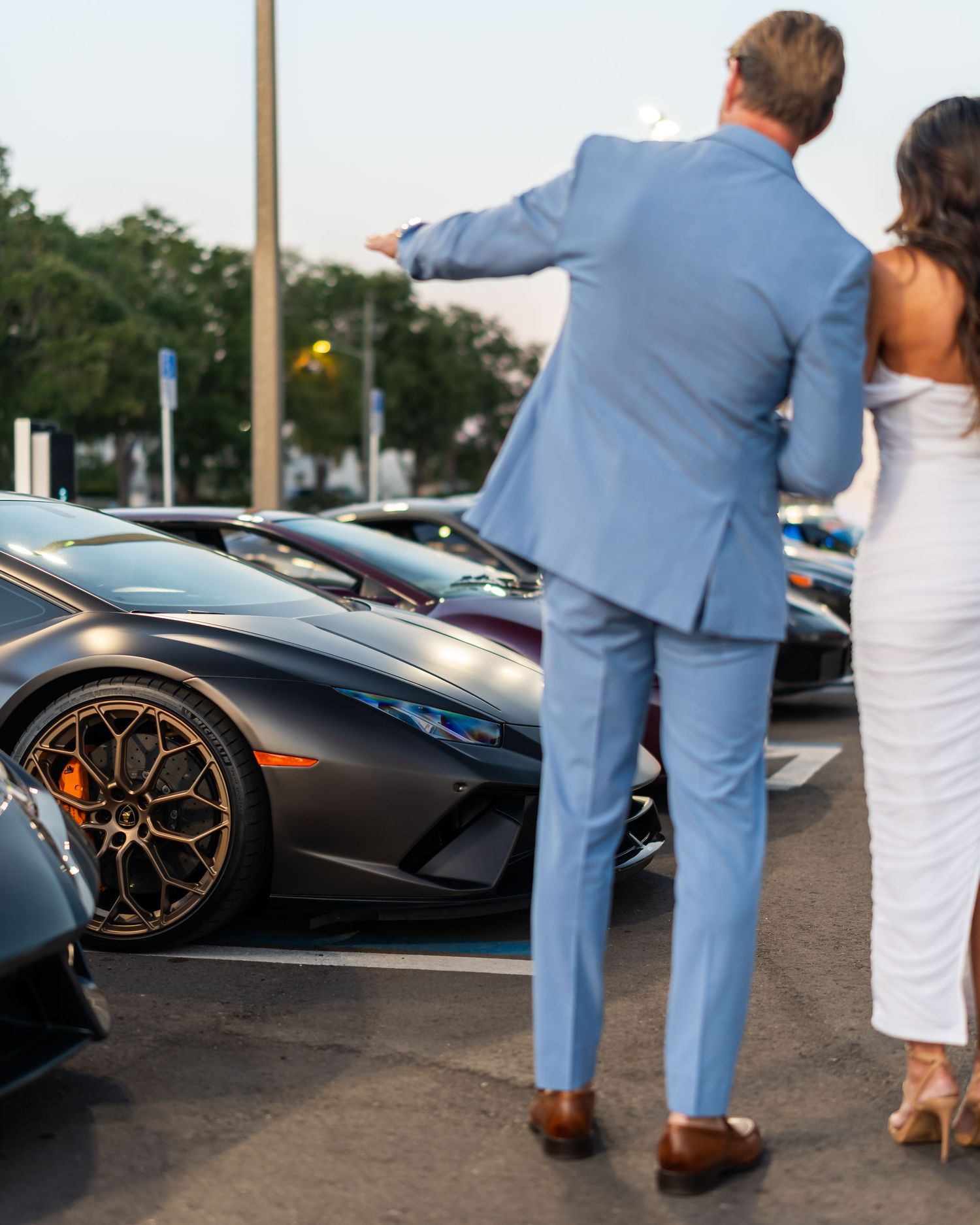 Couple dressed in formalwear standing beside a black Lamborghini Huracán at an outdoor automotive event