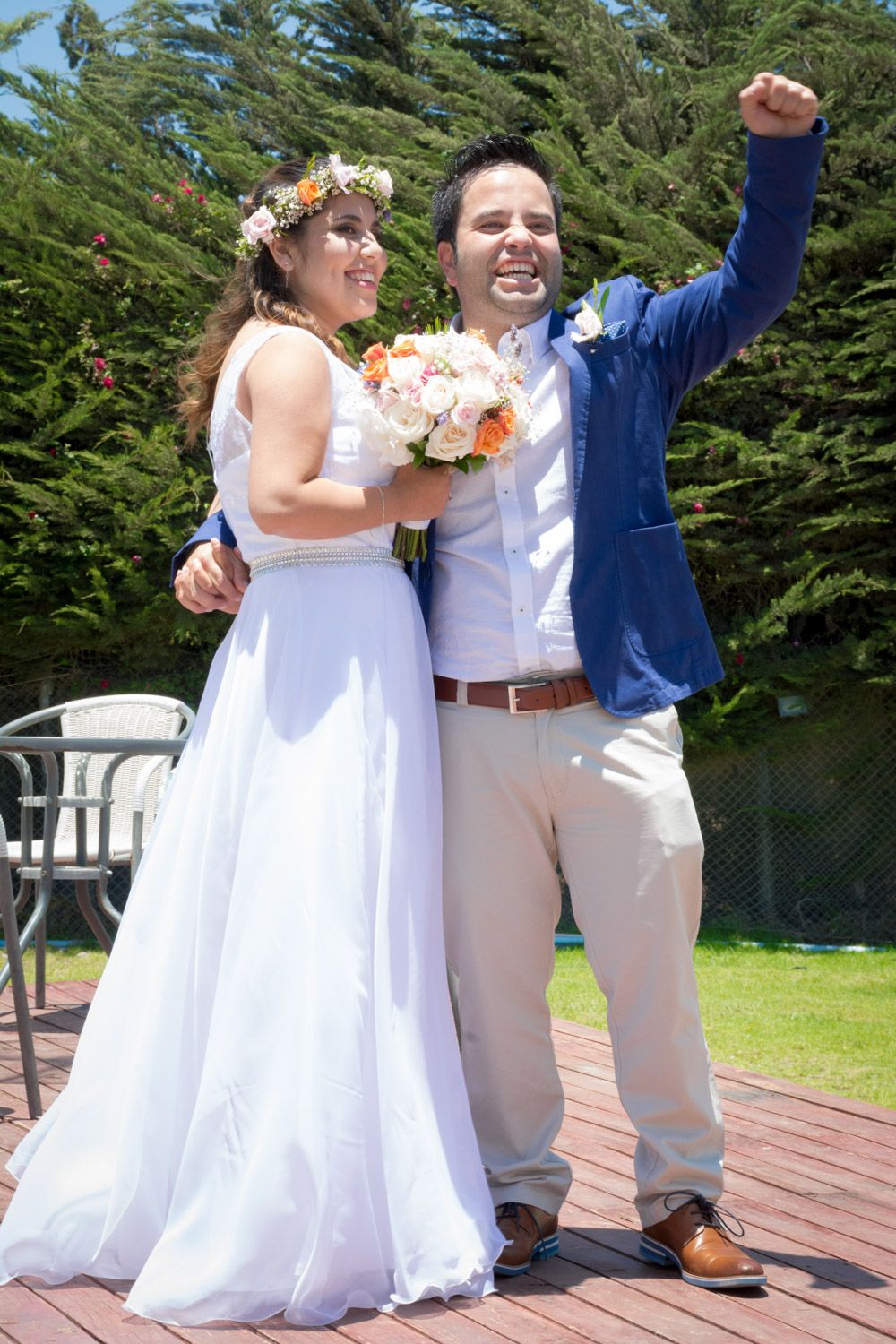 Joyful bride and groom celebrating outdoors, bride holding bouquet while groom raises fist triumphantly.