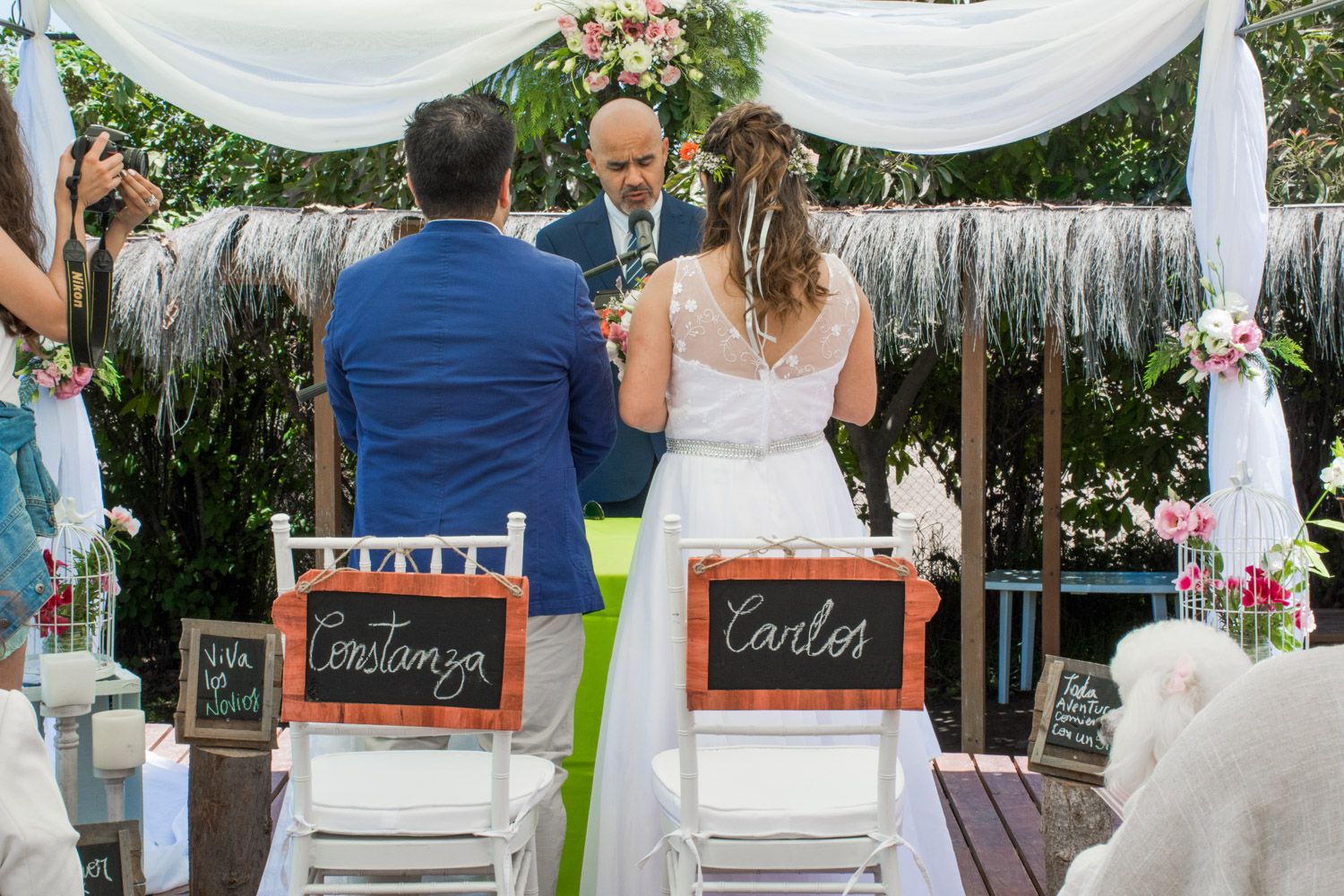 Bride and groom at outdoor wedding altar with personalized chalkboard name signs on white chairs.