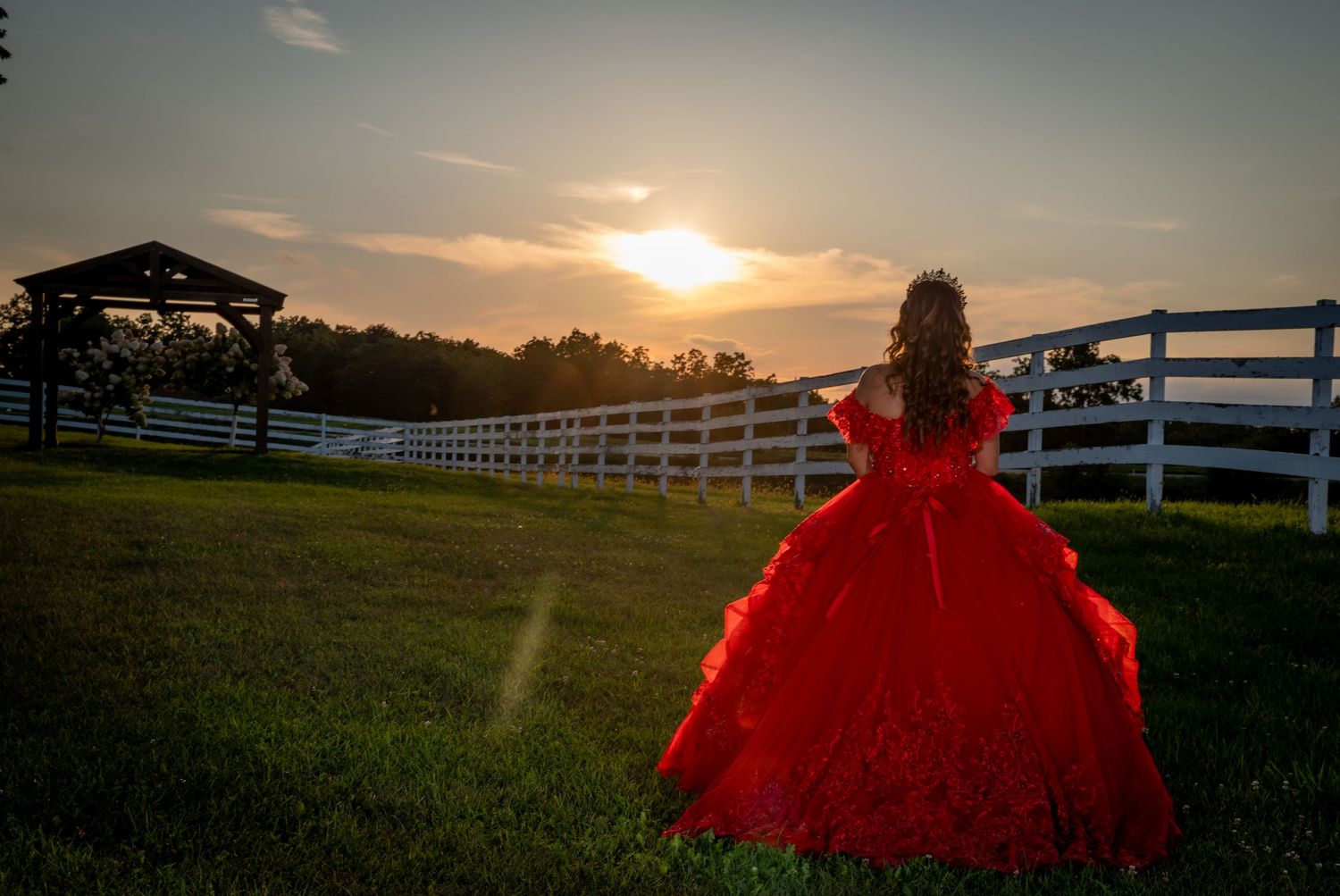 A girl in a red quinceañera ball gown stands by a white fence at sunset on a lush green farm.