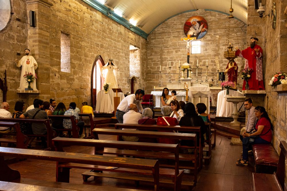 People praying inside a historic stone Catholic church with wooden pews, altar, and religious statues.