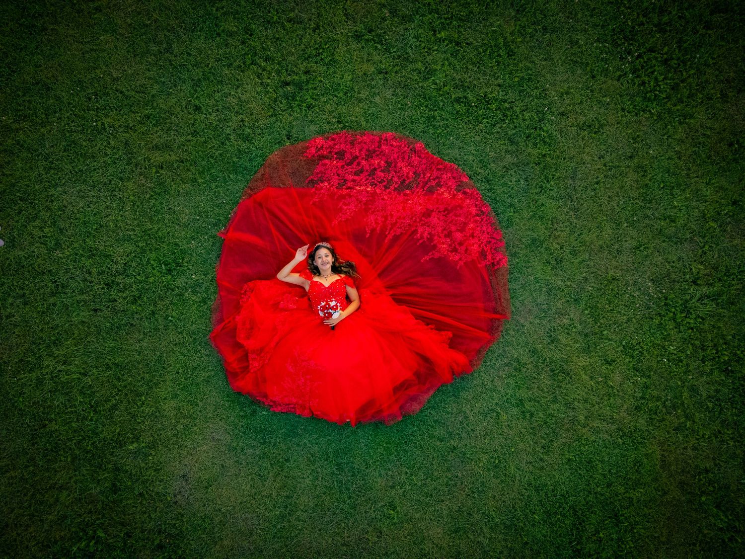 Aerial drone view of a quinceañera girl lying in the center of her spread red ball gown on green grass.