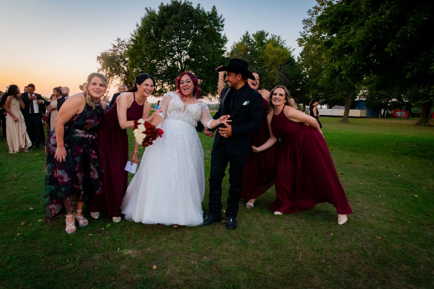 Joyful bride in white gown posing with bridesmaids in burgundy dresses during sunset wedding celebration.