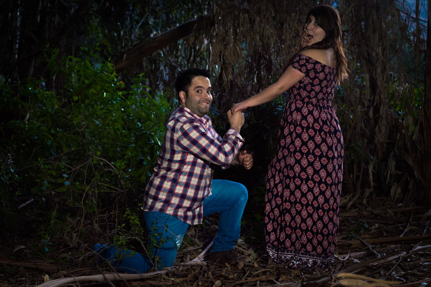 Man kneeling and holding womans hand in a dramatic romantic pose surrounded by trees and brush at night.