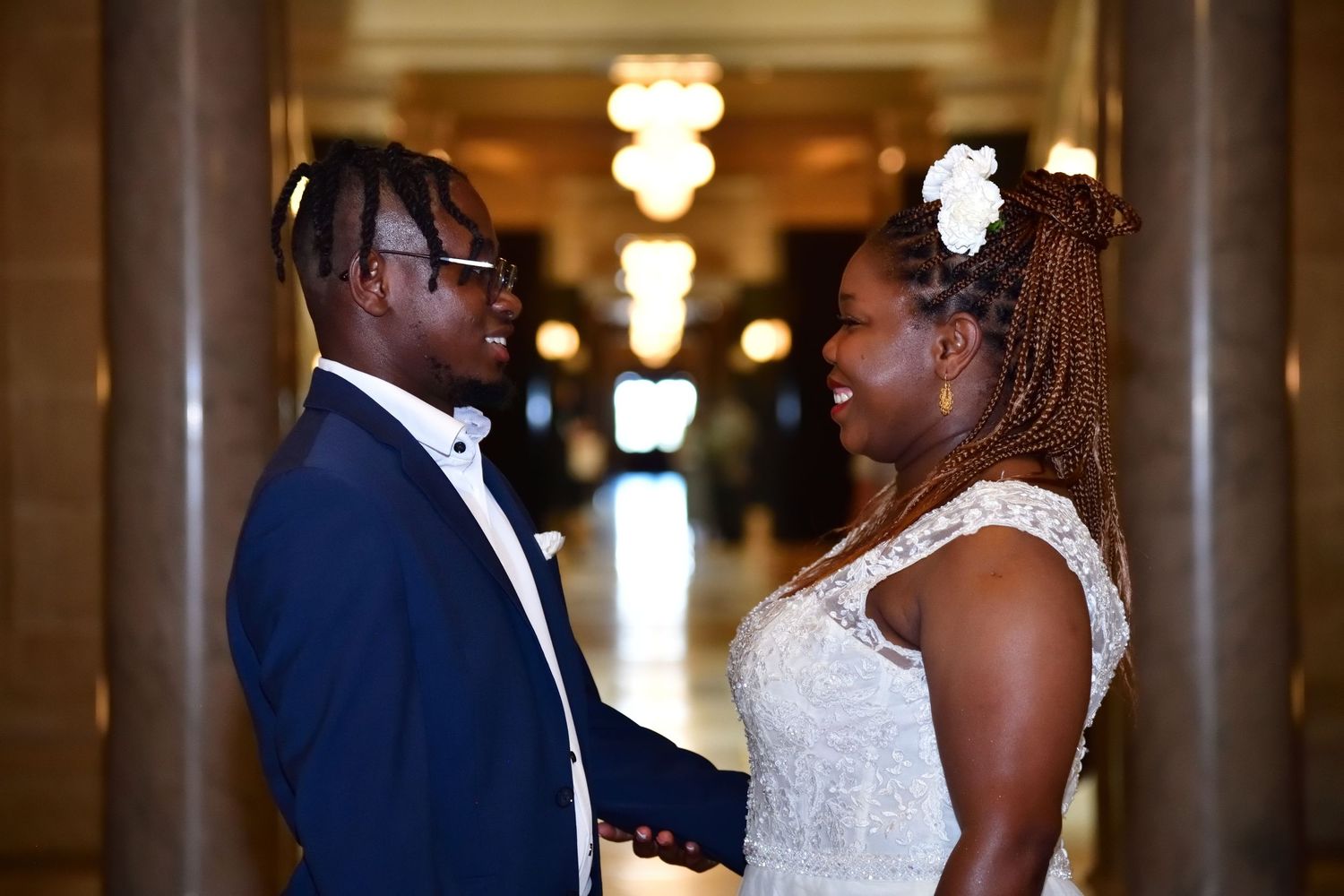 A joyful Black couple in wedding attire holds hands and smiles at each other in an elegant indoor venue.