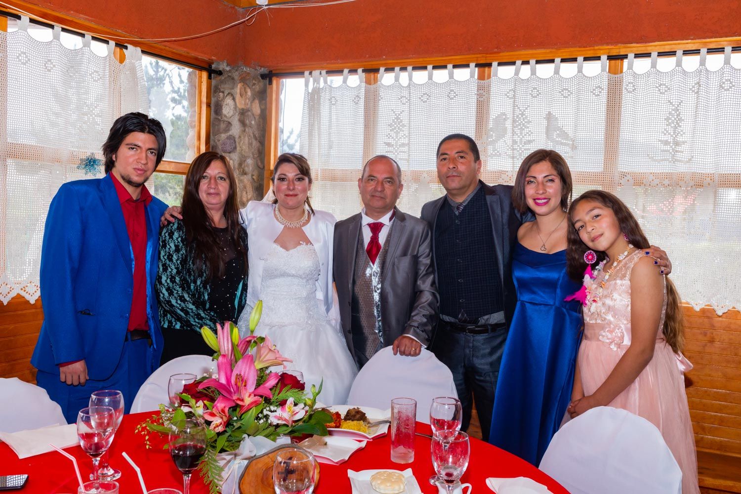 Wedding party group photo with bride, groom, and family members at decorated reception table.