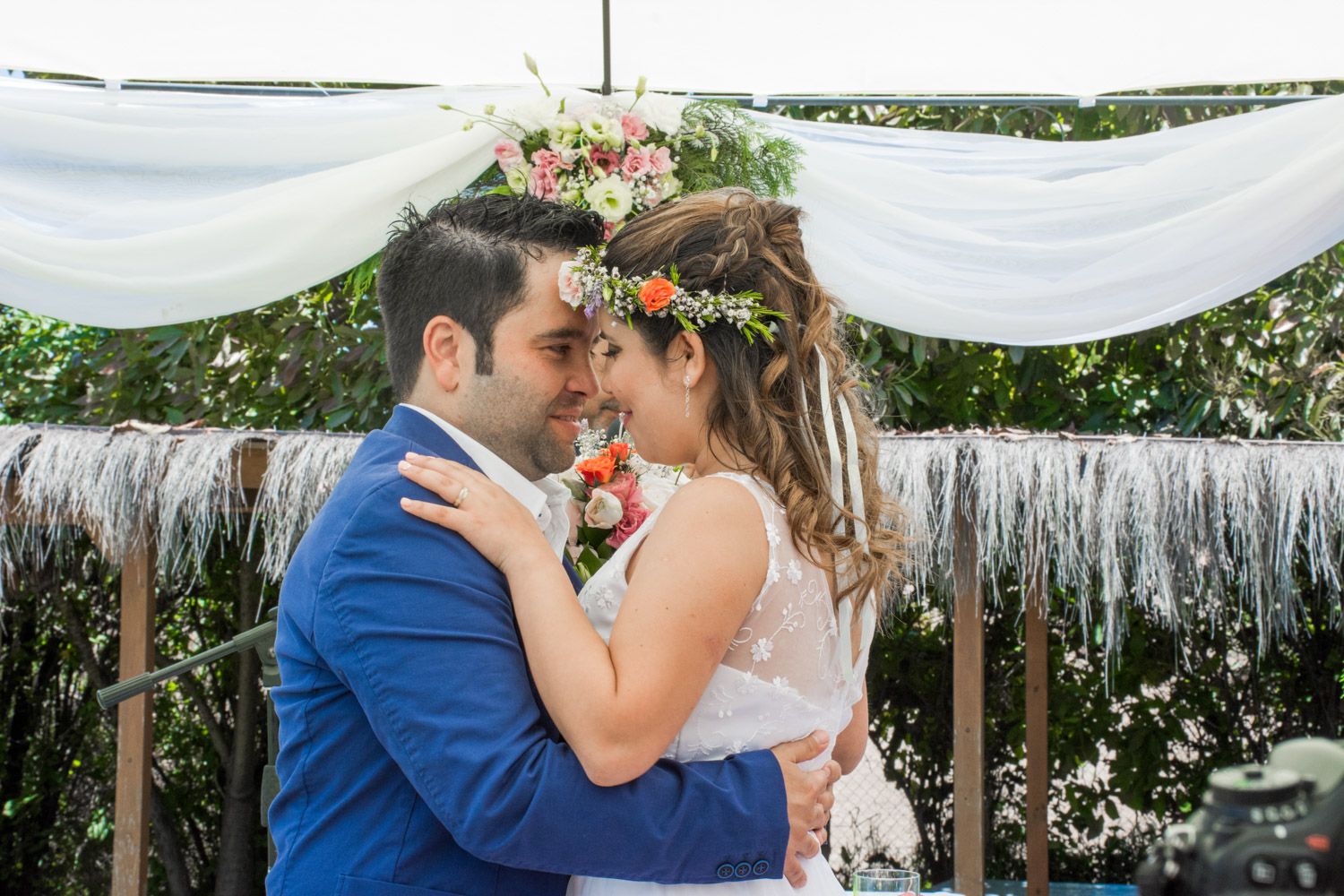 Bride wearing floral crown and groom in blue suit sharing romantic forehead touch at outdoor wedding ceremony.