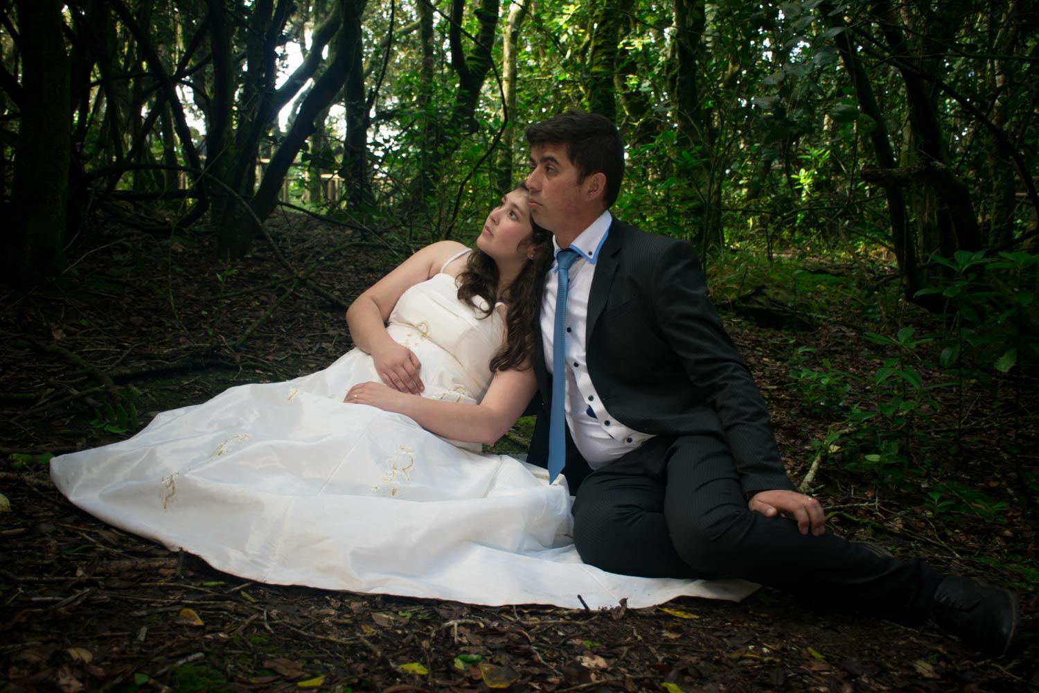 Newlywed couple sitting together on forest ground, bride in white gown, groom in dark suit with blue tie.