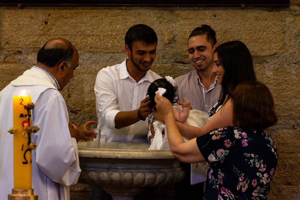Catholic priest performing a baptism ceremony with family gathered around a stone baptismal font in a church.