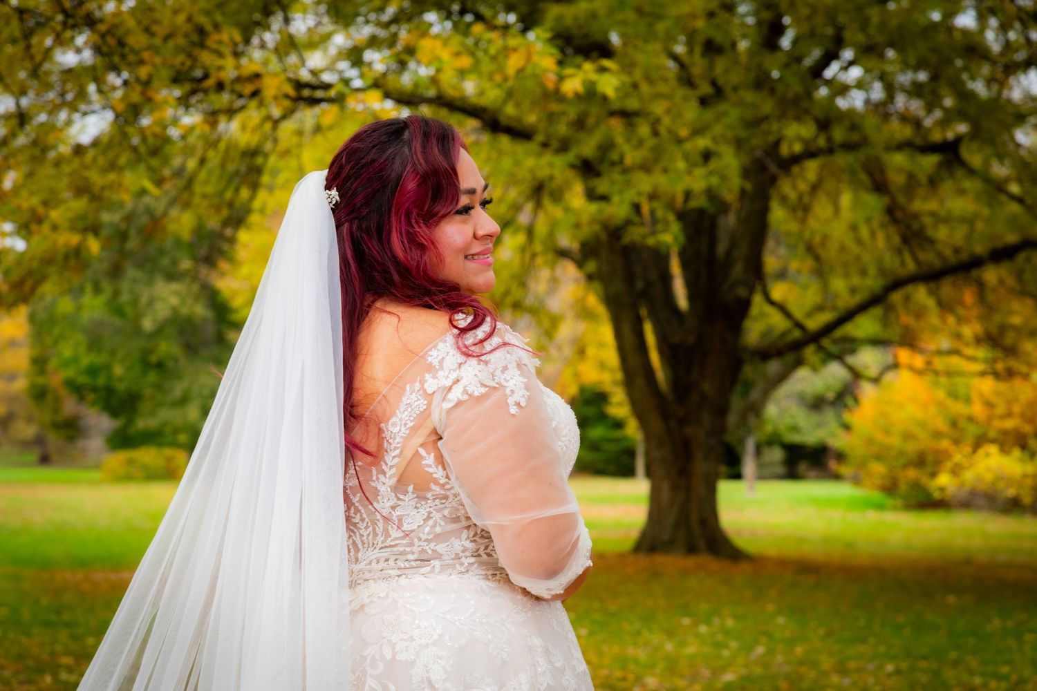Bride in white lace gown and veil poses outdoors among vibrant autumn foliage trees in fall wedding portrait.