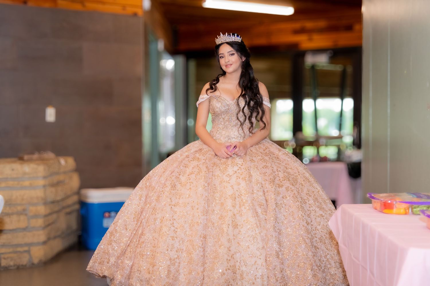 Teen girl in elegant rose gold quinceañera gown wearing a tiara stands in a decorated celebration venue.