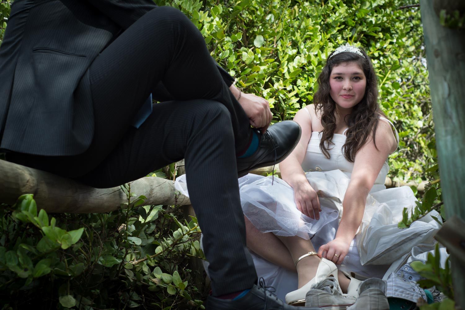 Bride adjusting white heels while groom ties shoes on rustic wooden fence outdoors.