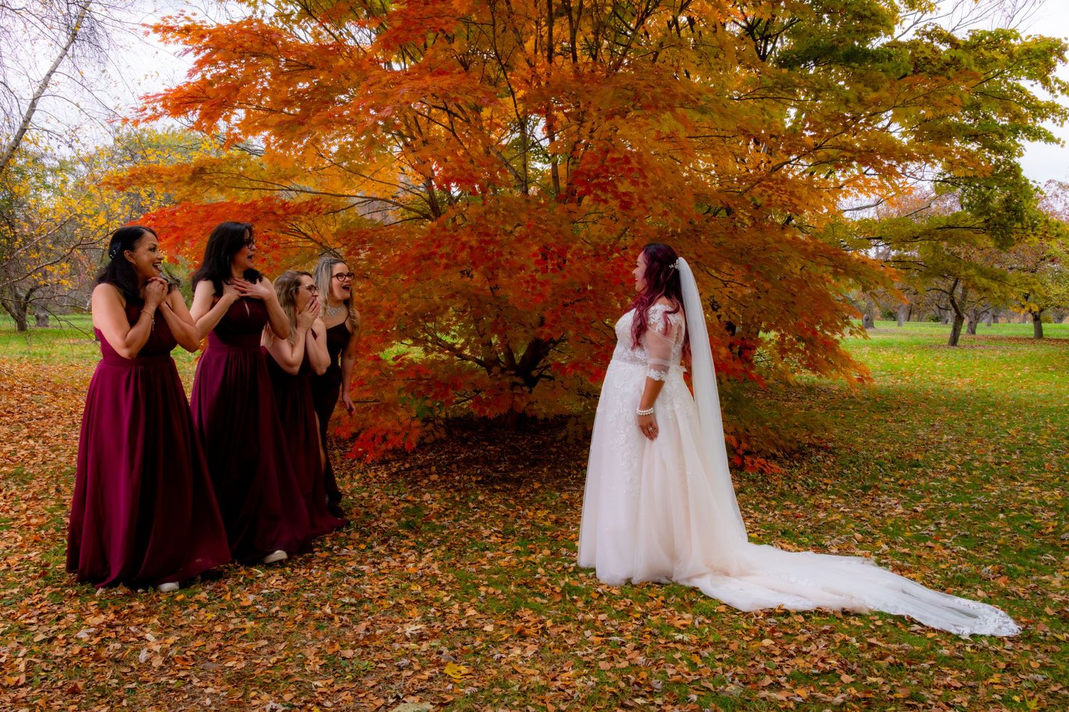 Bridesmaids in burgundy dresses photograph bride in white gown amid vibrant fall foliage.