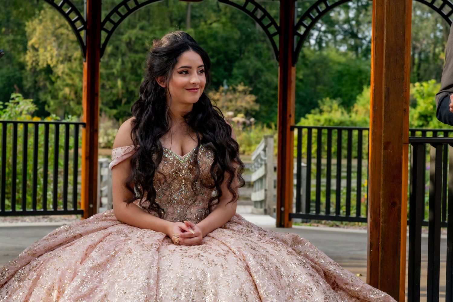 Young woman in rose gold quinceañera ballgown poses elegantly in a wooden gazebo surrounded by greenery.