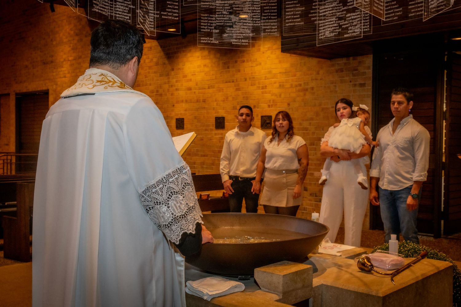 Priest performing a baptism ceremony at a church font with godparents and baby dressed in white.