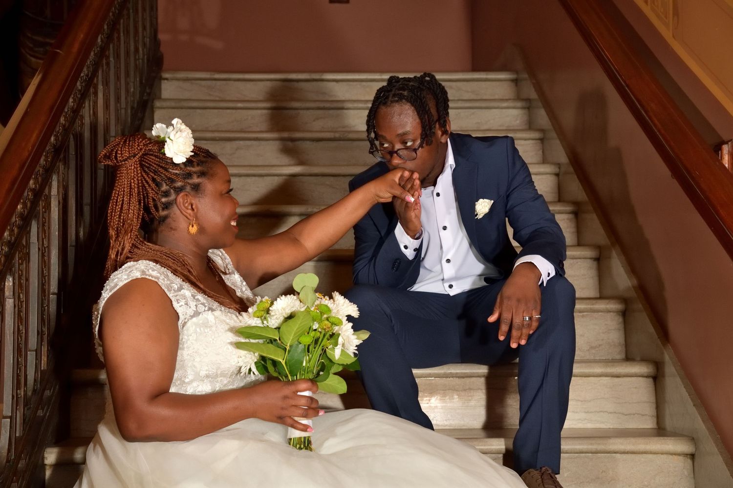 A bride and groom sit on marble stairs sharing a tender moment with a green and white bridal bouquet.