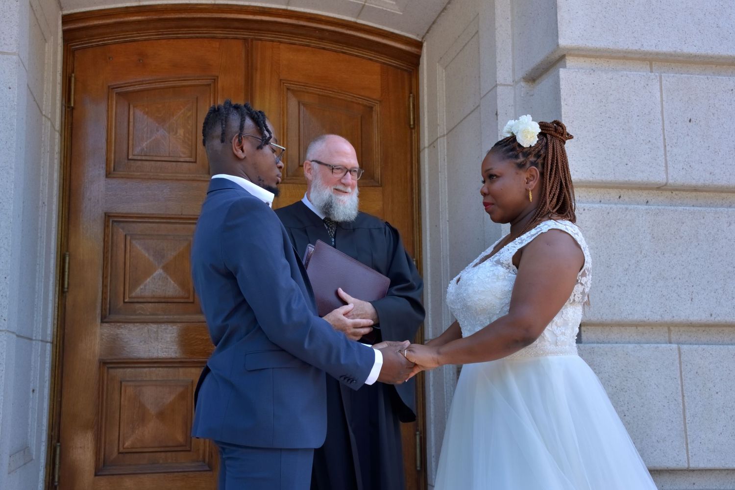Couple exchanges vows before officiant at wooden church doors during outdoor wedding ceremony.