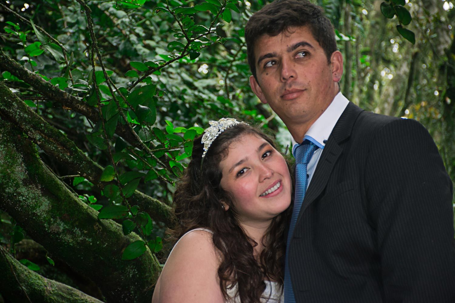 Happy wedding couple posing together against a moss-covered tree in a lush green forest setting.