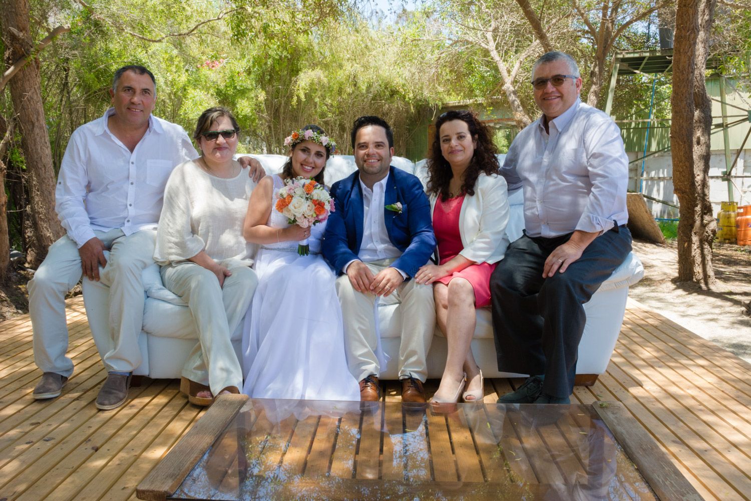 Wedding party of six people posing together on a white outdoor sofa surrounded by trees.