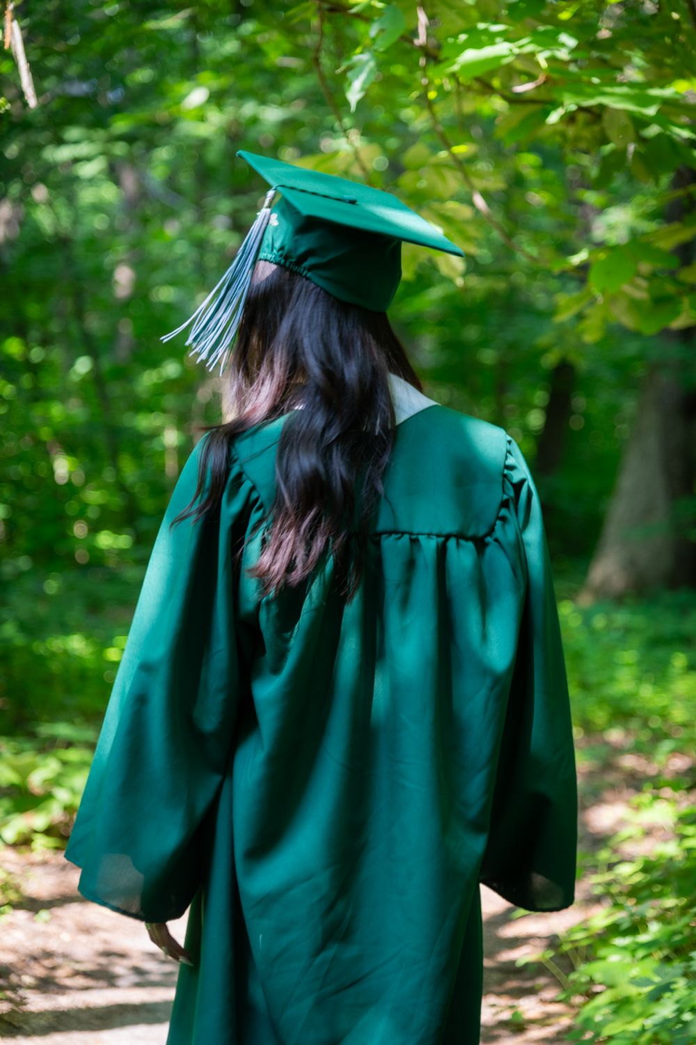 Young woman in green graduation cap and gown walking through lush forest path from behind.