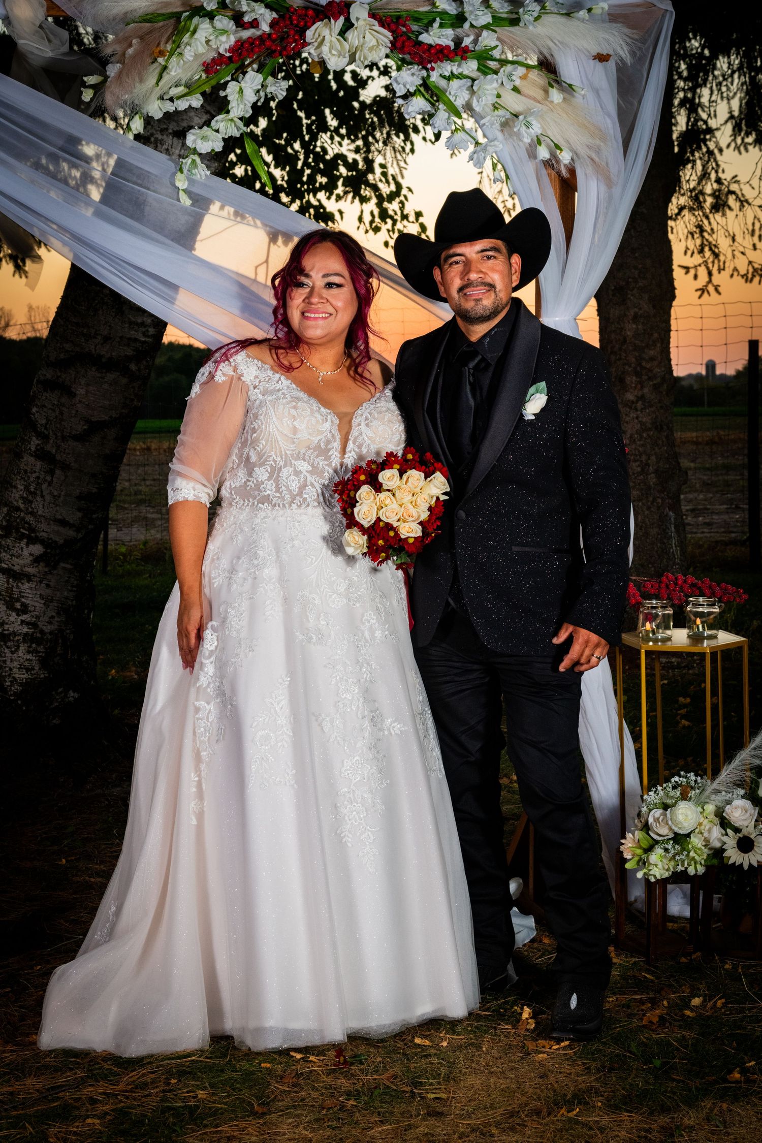 Bride in lace gown and groom in black cowboy hat posing under floral wedding arch at sunset.
