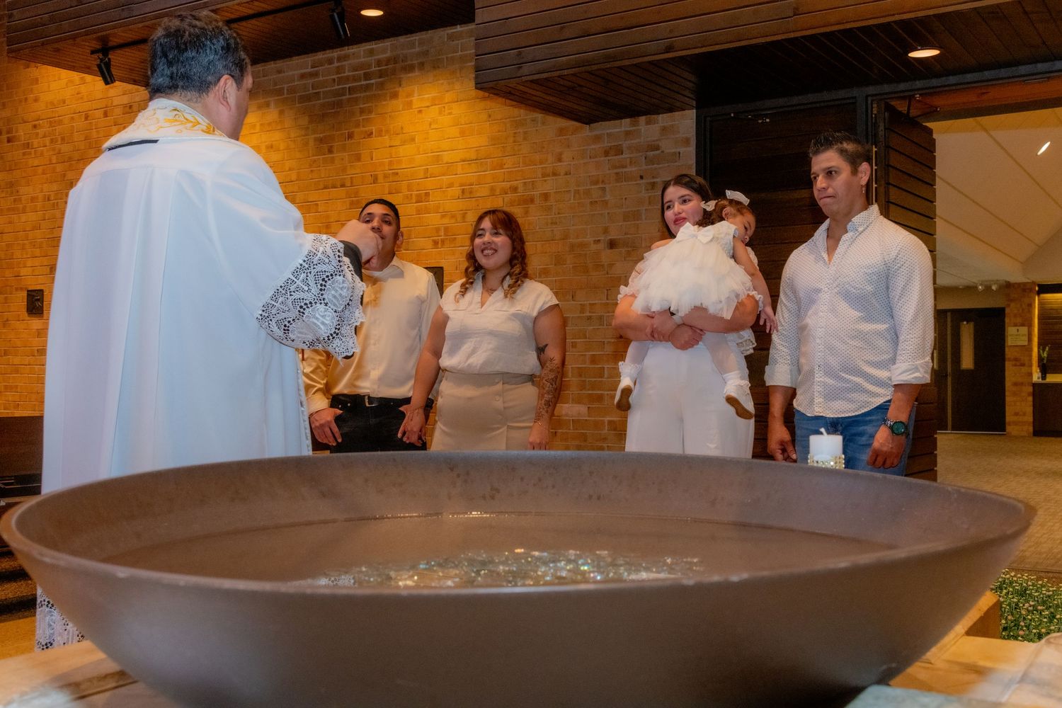 A priest performs a baptism ceremony over a large brown font with family and baby present in a church.