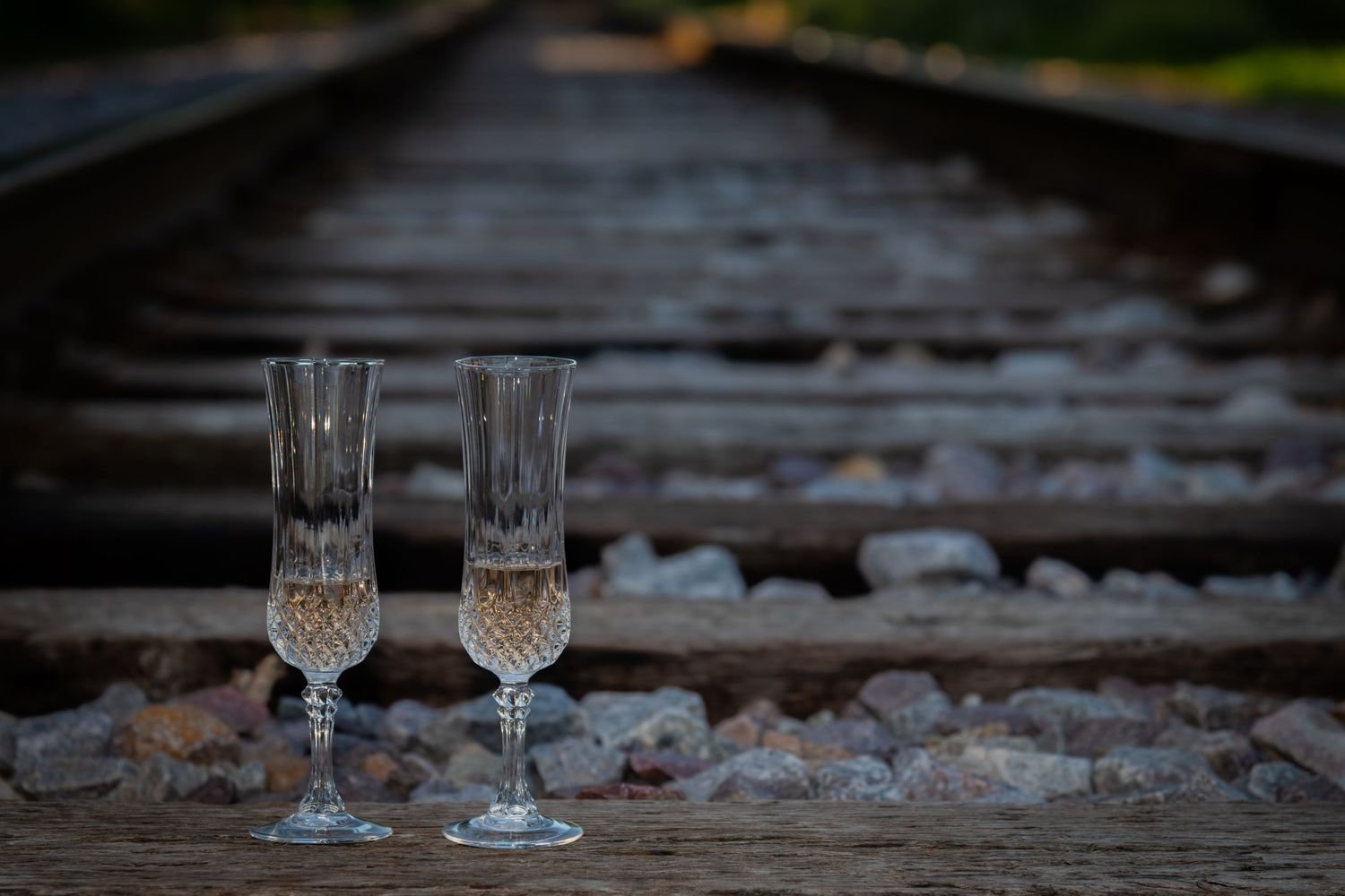 Two crystal champagne flutes filled with sparkling wine rest on vintage railroad tracks at dusk.
