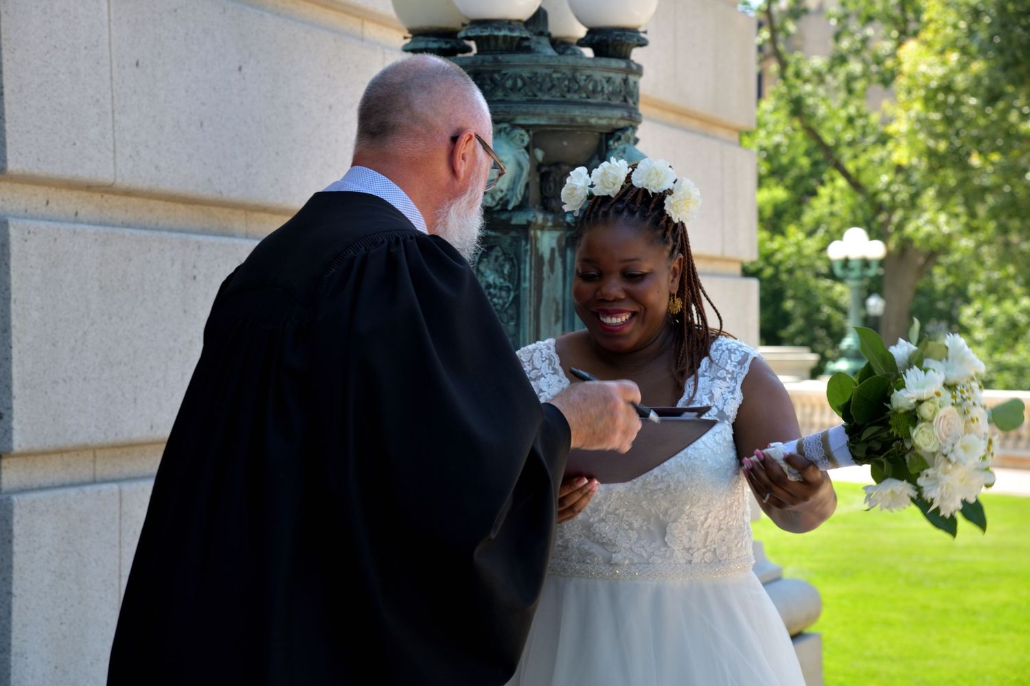 Bride in white gown laughs with officiant in black robes during outdoor wedding celebration.