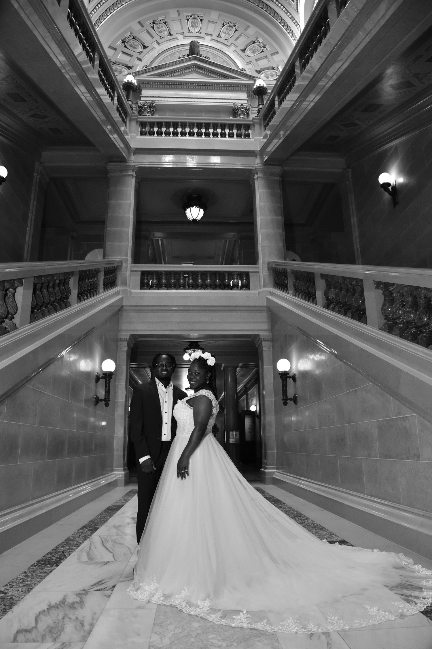 Black and white photo of a bride and groom standing beneath an ornate grand staircase in a historic building.