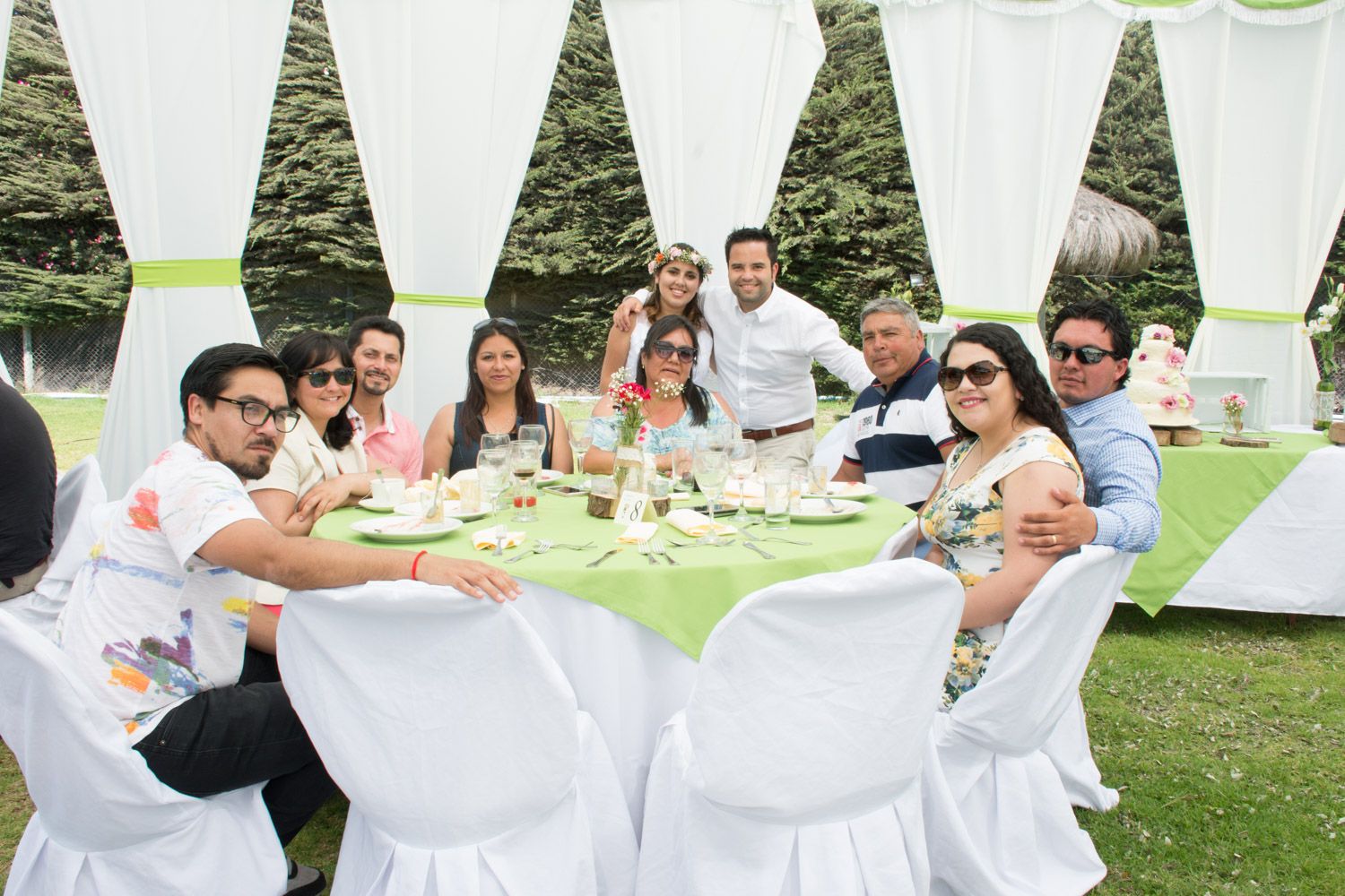 Wedding reception guests gathered around a decorated green and white table outdoors.