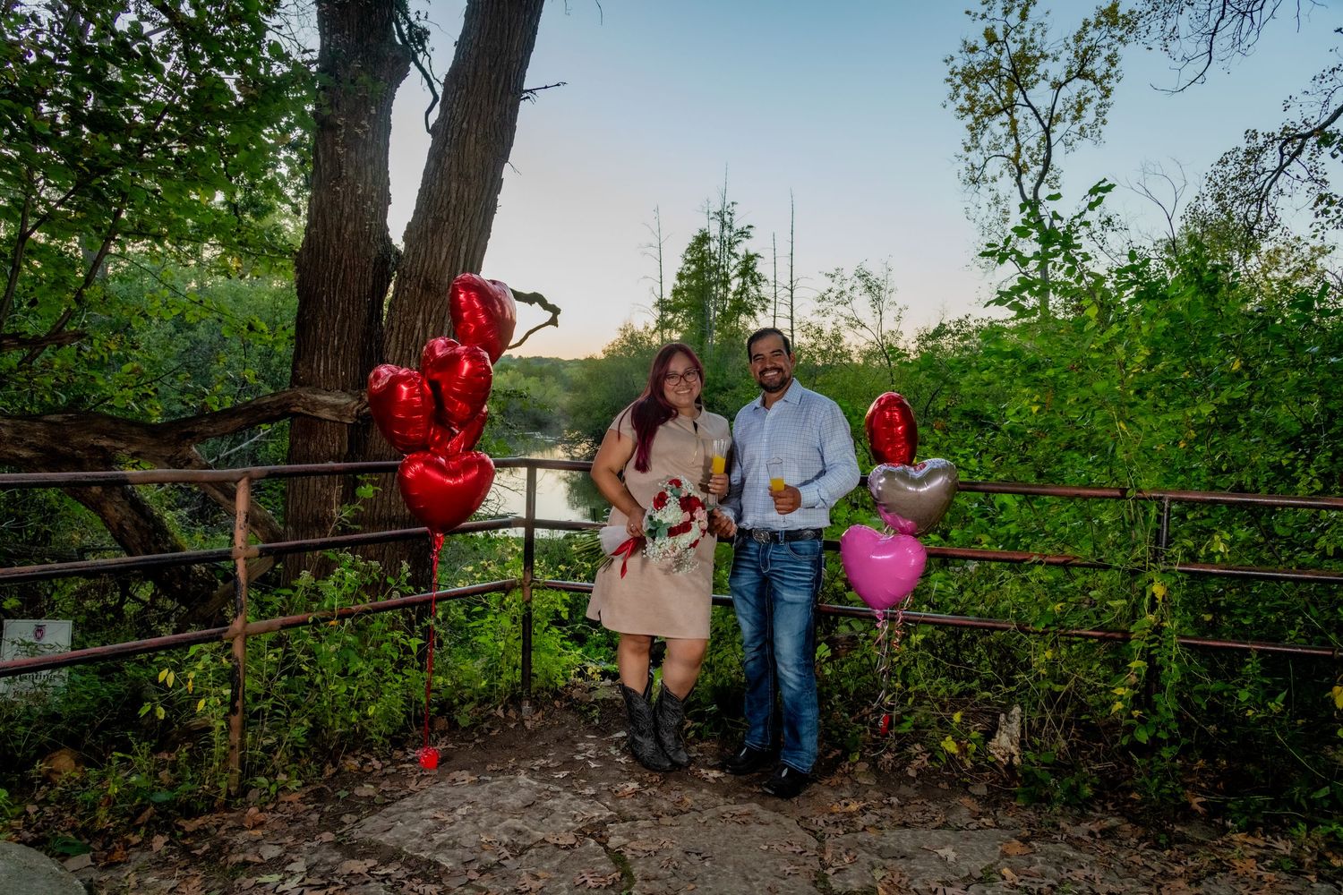 Couple posing with red heart balloons and bouquet at an outdoor riverside engagement celebration.