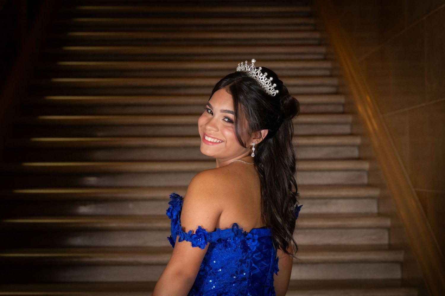 Young woman in royal blue quinceañera gown and tiara smiling on elegant staircase.