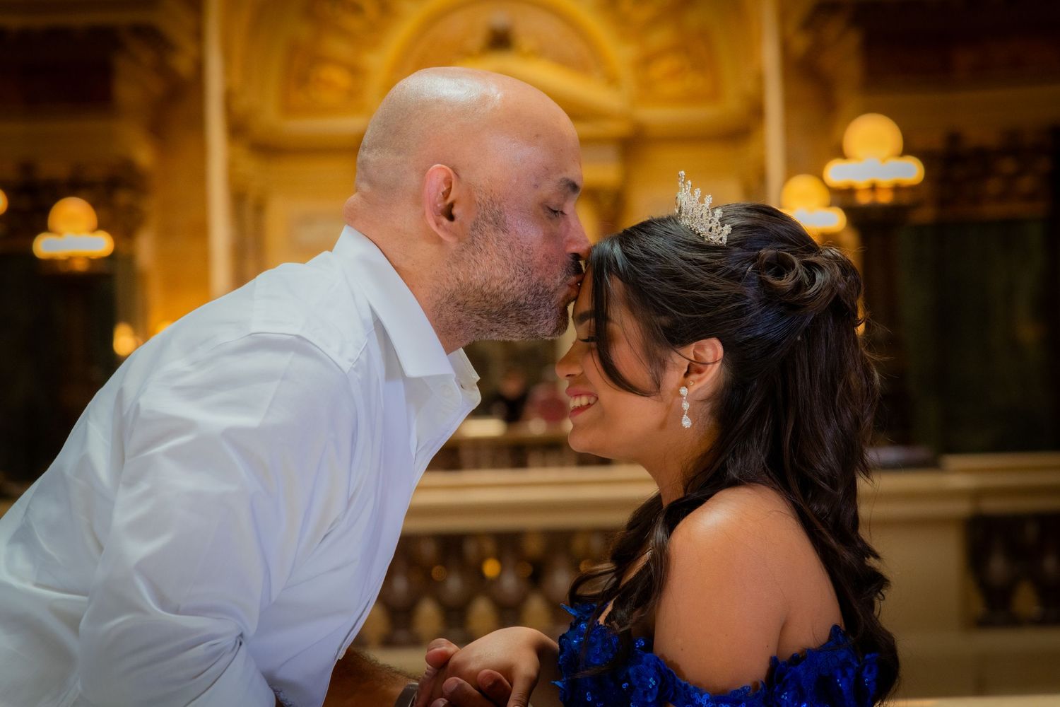 A bald groom leans in to kiss his bride wearing a tiara and blue gown in an ornate golden venue.