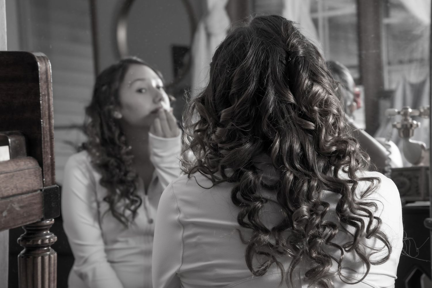 A woman with curly hair applies makeup while looking into a mirror in a black and white photo.