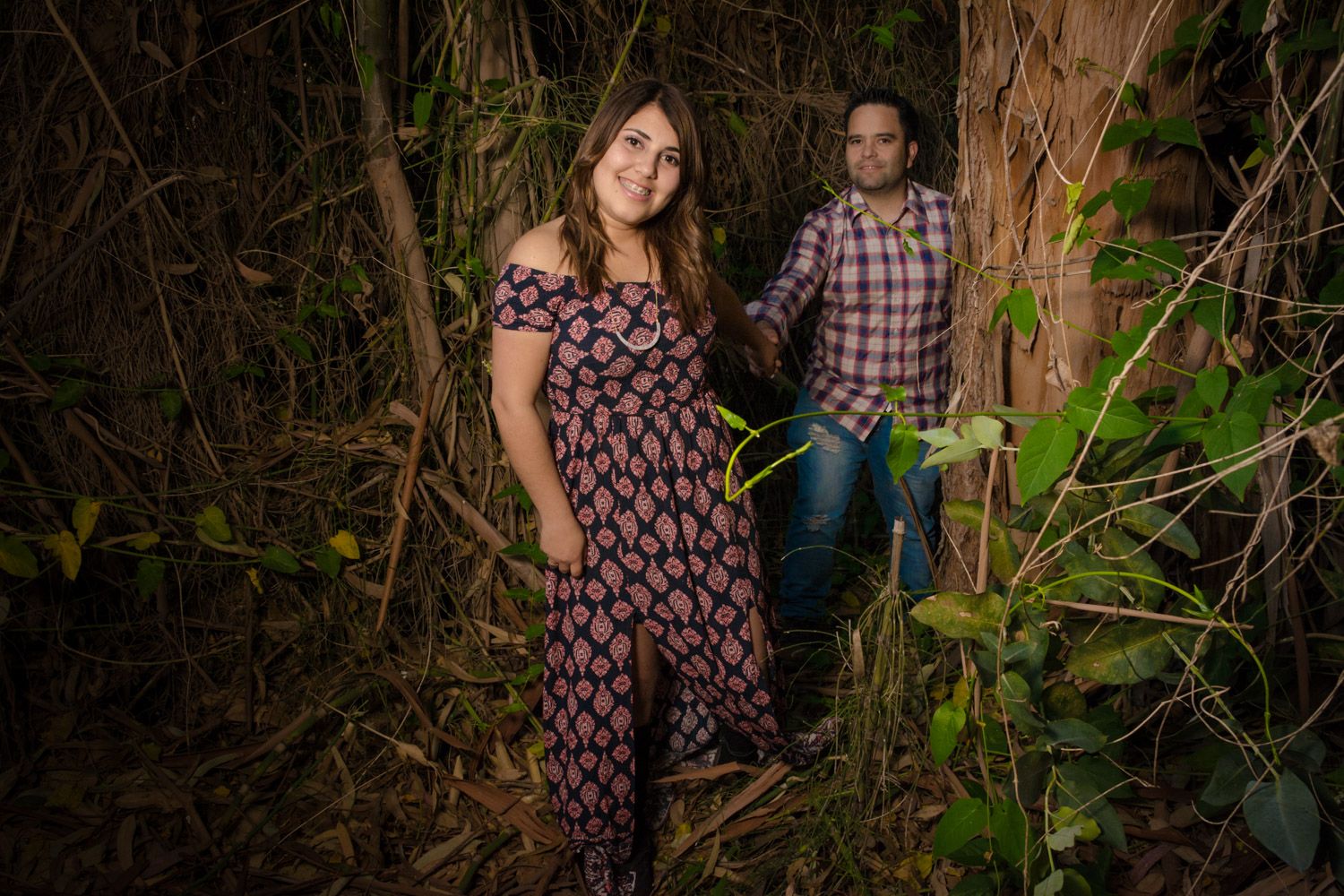 Couple posing among dense tropical vines and foliage at night, woman in floral maxi dress smiling forward.